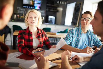Group of young business professionals discussing ideas while collaborating on startup project during meeting in modern office, focus on blonde young woman pointing at document