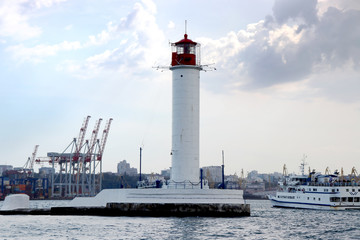 Fototapeta na wymiar Lighthouse in the sea against the backdrop of a cargo port. Summer seascape with a white lighthouse with red top. Black Sea. Sea Port of Odessa.