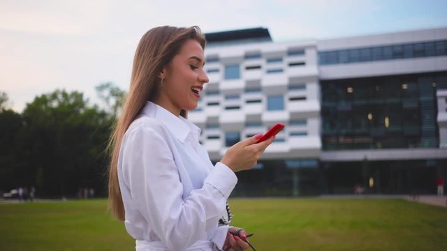 Pleasant-looking Caucasian girl walking through the street holding red smartphone in hand, smiling. Beautiful young Ukrainian woman chewing bubble gum. Outdoors.