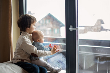 Preschool boy, holding his baby brother, sitting by the window in living room, looking at a snowy...