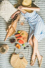 Photo sur Plexiglas Pique-nique Cadre de pique-nique d& 39 été. Femme en robe à rayures en lin et chapeau de paille assis avec un verre de vin rose à la main, des fruits frais et de la baguette sur une couverture, vue de dessus. Concept de rassemblement ou de déjeuner en plein air