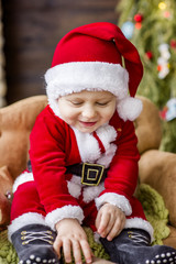 Close-up portrait of a Little boy dressed as a red Santa Claus, playing by a stone fireplace with bright garlands, putting a gift box under the Christmas tree. New Year card. The baby laughs, has fun.
