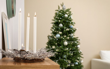 Burning candles with wreath on table and Christmas tree in stylish living room interior