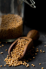 Heap of raw, unprocessed mustard seed kernels in glass jar and wooden scoop on black table