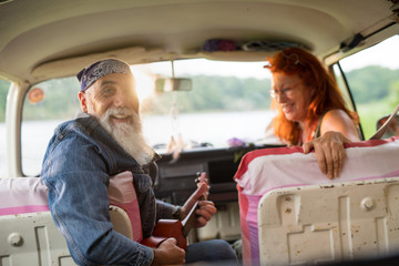 An old hipster couple sitting in a van, the man playing ukulele 