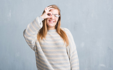 Young adult woman over grey grunge wall wearing glasses with happy face smiling doing ok sign with hand on eye looking through fingers