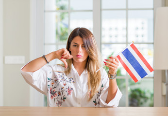 Young woman at home holding flag of Thailand with angry face, negative sign showing dislike with...