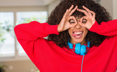African american woman wearing headphones doing ok gesture like binoculars sticking tongue out, eyes looking through fingers. Crazy expression.