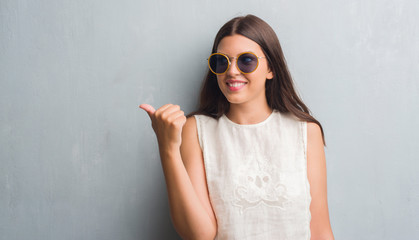 Young brunette woman over grunge grey wall wearing retro sunglasses pointing and showing with thumb up to the side with happy face smiling
