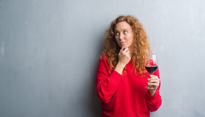 Young redhead woman over grey grunge wall drinking a glass of wine serious face thinking about...