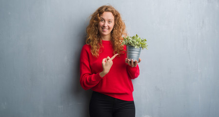 Young redhead woman over grey grunge wall holding plant pot very happy pointing with hand and finger