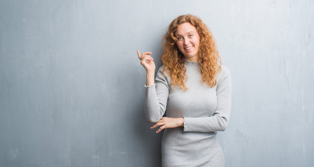 Young redhead woman over grey grunge wall very happy pointing with hand and finger to the side