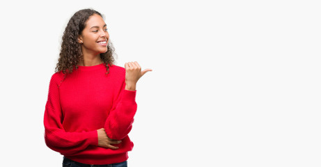 Young hispanic woman wearing red sweater smiling with happy face looking and pointing to the side...