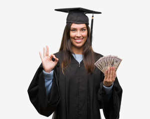 Young hispanic woman wearing graduated uniform holding dollars doing ok sign with fingers,...