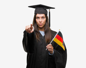Young hispanic woman wearing graduated uniform holding flag of germany pointing with finger to the camera and to you, hand sign, positive and confident gesture from the front