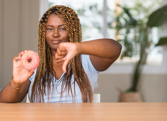 African american woman eating doughnut with angry face, negative sign showing dislike with thumbs...