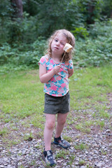 Little blond Caucasian girl is eating a marshmallow on the stick. She is in the park. She is smiling.