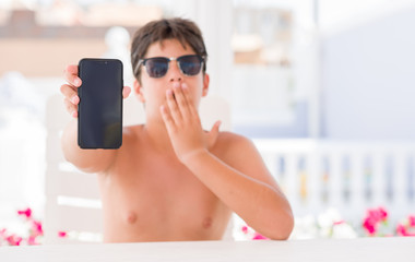 Young child on holidays showing smartphone cover mouth with hand shocked with shame for mistake, expression of fear, scared in silence, secret concept