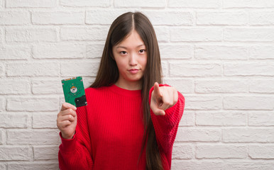 Young Chinese woman over brick wall holding hard drive pointing with finger to the camera and to you, hand sign, positive and confident gesture from the front