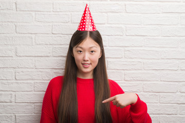Young Chinese woman over brick wall wearing birthday hat with surprise face pointing finger to...