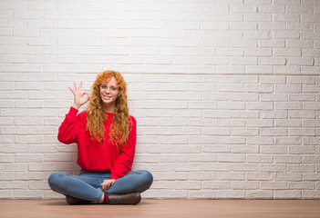 Young redhead woman sitting over brick wall smiling positive doing ok sign with hand and fingers. Successful expression.