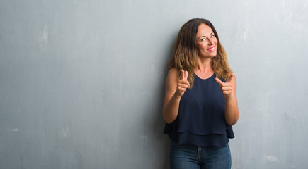 Middle age hispanic woman standing over grey grunge wall pointing fingers to camera with happy and...