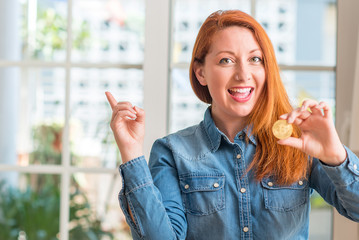 Redhead woman holding bitcoin cryptocurrency at home very happy pointing with hand and finger to...