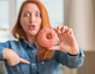 Redhead woman holding donut at home with surprise face pointing finger to himself