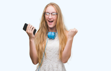 Blonde teenager woman holding smartphone and wearing headphones screaming proud and celebrating victory and success very excited, cheering emotion