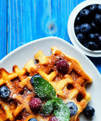 Traditional belgian waffles with fresh berrys and sugar powder on white plate, blue wooden background. Flat lay, top view, copy space.
