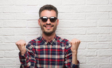 Young adult man wearing sunglasses standing over white brick wall screaming proud and celebrating victory and success very excited, cheering emotion