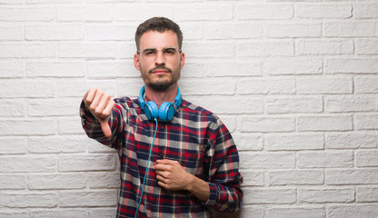 Young adult man over brick wall wearing headphones with angry face, negative sign showing dislike with thumbs down, rejection concept