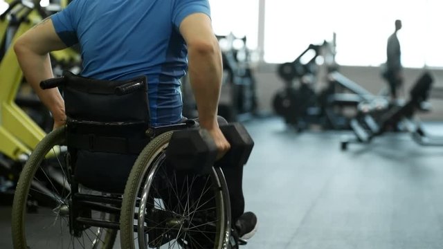 Tilt up with rear view of unrecognizable handicapped man in wheelchair doing one-arm dumbbell rows in gym