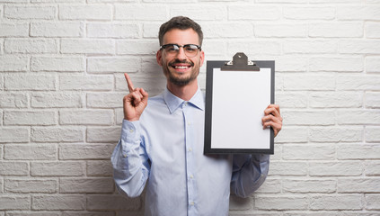 Young adult business man over brick wall holding clipboard surprised with an idea or question...