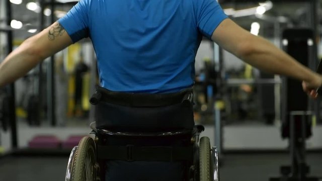 Tilt up with rear view of unrecognizable paraplegic man in wheelchair doing cable chest fly exercise in gym