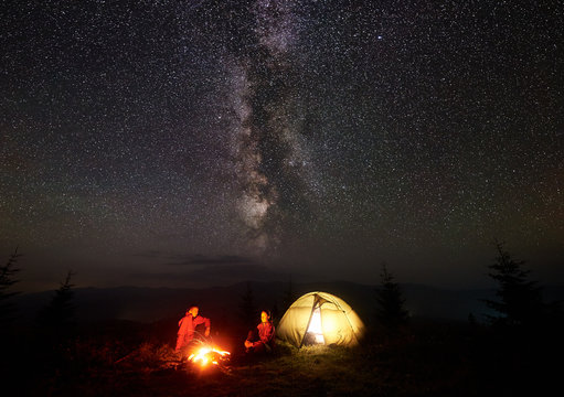 Romantic couple hikers man and woman having a rest on grassy valley near illuminated tent, burning campfire under starry sky and Milky way, enjoying quiet night camping in mountains