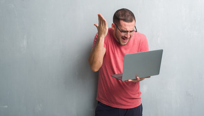 Young caucasian man over grey grunge wall using computer laptop annoyed and frustrated shouting with anger, crazy and yelling with raised hand, anger concept