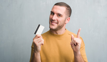 Young caucasian man over grey grunge wall holding credit card surprised with an idea or question...