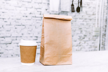 Cup of coffee and blank paper bag with copy space for your brand on table against white brick wall background