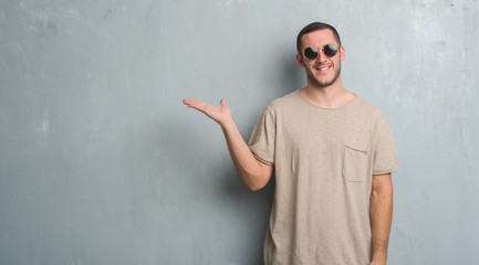 Young caucasian man over grey grunge wall wearing sunglasses smiling cheerful presenting and pointing with palm of hand looking at the camera.