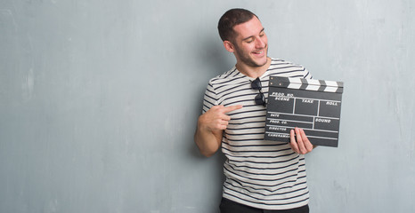 Young caucasian man over grey grunge wall holding movie clapboard very happy pointing with hand and...