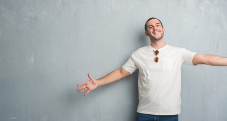 Young caucasian man over grey grunge wall wearing sunglasses looking at the camera smiling with open arms for hug. Cheerful expression embracing happiness.