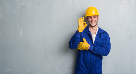 Young caucasian man over grey grunge wall wearing contractor uniform and safety helmet doing ok sign with fingers, excellent symbol