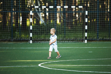 Fototapeta premium Happy cute blond boy in blue t-shirt and shorts on a football field without a ball.