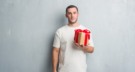 Young caucasian man over grey grunge wall holding a present with a confident expression on smart face thinking serious