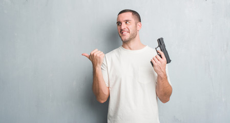 Young caucasian criminal man over grey grunge wall holding gun pointing and showing with thumb up...