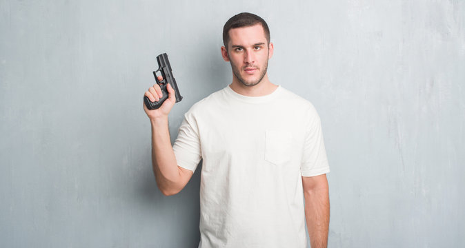 Young caucasian criminal man over grey grunge wall holding gun with a confident expression on smart face thinking serious