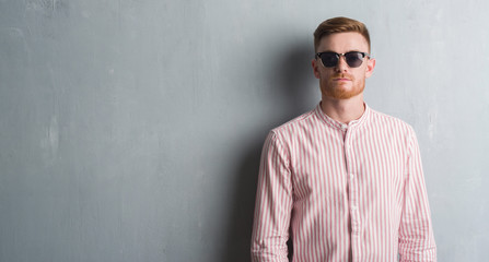 Obraz premium Young redhead man over grey grunge wall wearing sunglasses with a confident expression on smart face thinking serious