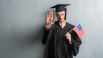 Young redhead man over grey grunge wall wearing graduate uniform holding america flag with open...