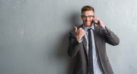 Young redhead  business man over grey grunge wall talking on the phone pointing and showing with...